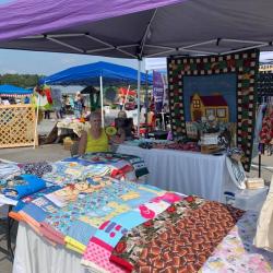 A market stall with patchwork quilts, fabrics, and a decorative panel, a woman is sitting at the table