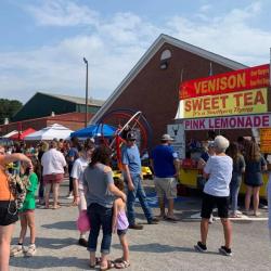 An outdoor fair — a crowd gathered around a vendor booth selling "Venison, Sweet Tea, Pink Lemonade"