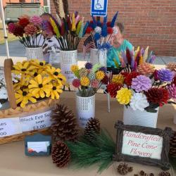 A booth with brightly painted pine cones arranged as flowers, and an elderly woman sitting behind the counter.